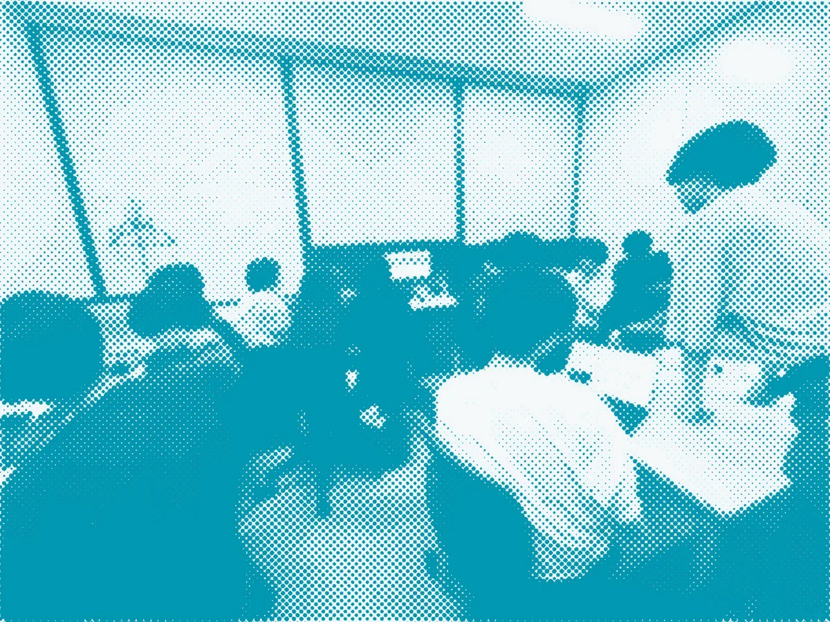 部内ハッカソンの発表会。机を囲んで発表を聞くメンバーと、立って説明するメンバー(網点加工)